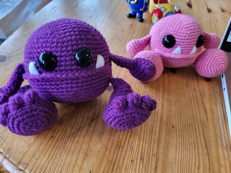 May include: Two crocheted plush toys, one purple and one pink, with white eyes and teeth. The purple toy has long arms and legs, while the pink toy has shorter arms and legs.