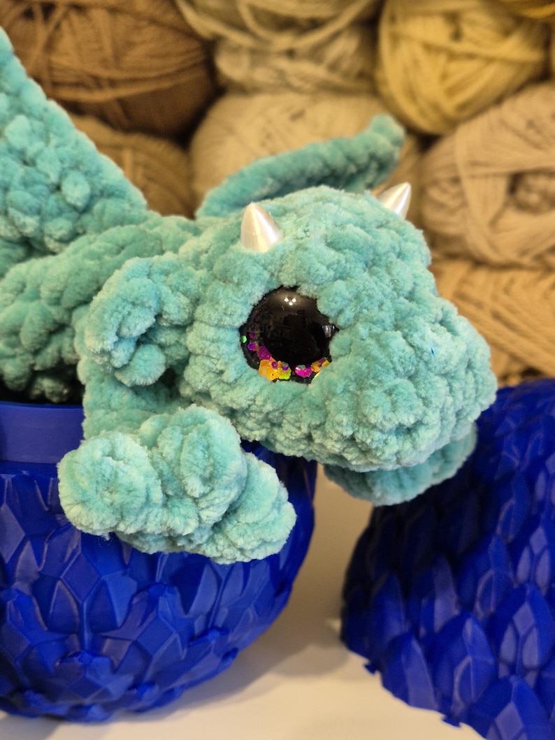May include: A teal blue crocheted dragon with large googly eyes, sitting inside a blue dragon egg. The egg is made of plastic and has a scaly texture.