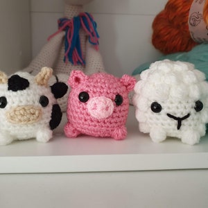 May include: Three crocheted stuffed animals: a cow, a pig, and a sheep. The cow is black and white, the pig is pink, and the sheep is white. All three animals have black eyes and are sitting on a white shelf.