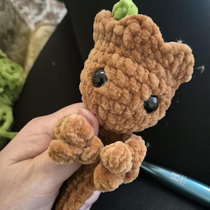 May include: A small, brown, crocheted plush toy shaped like a tree character. It features a textured, fuzzy yarn surface, two large, round, black plastic eyes, and a small, bright green sprout emerging from the top of its head.