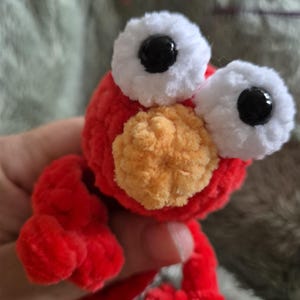 May include: A small, handmade crochet plush toy featuring a bright red round head and body made from soft yarn. It has two large, prominent white circular eyes with black pupils and a textured, round orange nose centered on the face.