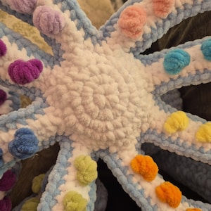 May include: A handmade, plush octopus toy in a vibrant color palette. The octopus features a white body, light blue tentacles, and colorful pom-poms in shades of purple, pink, blue, yellow, and orange. The soft, textured design is suitable for children.