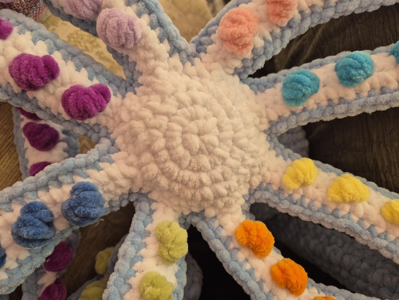 May include: A handmade, plush octopus toy in a vibrant color palette. The octopus features a white body, light blue tentacles, and colorful pom-poms in shades of purple, pink, blue, yellow, and orange. The soft, textured design is suitable for children.