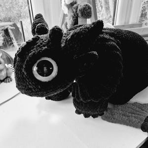 Peut inclure: Un grand jouet en forme de dragon noir crocheté, avec un œil rond proéminent. Le dragon présente des détails texturés et est posé sur une surface blanche. Un petit animal en peluche est visible en arrière-plan.