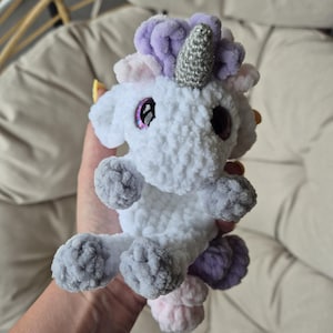 Puede incluir: Un unicornio de crochet blanco con una melena y cola de color morado y rosa. El unicornio tiene un cuerno de purpurina plateado y grandes ojos rosas.