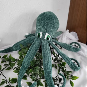 May include: A green crocheted octopus plush toy with white accents on the tentacles and large green eyes. The octopus is sitting on a white surface with green leafy plants.
