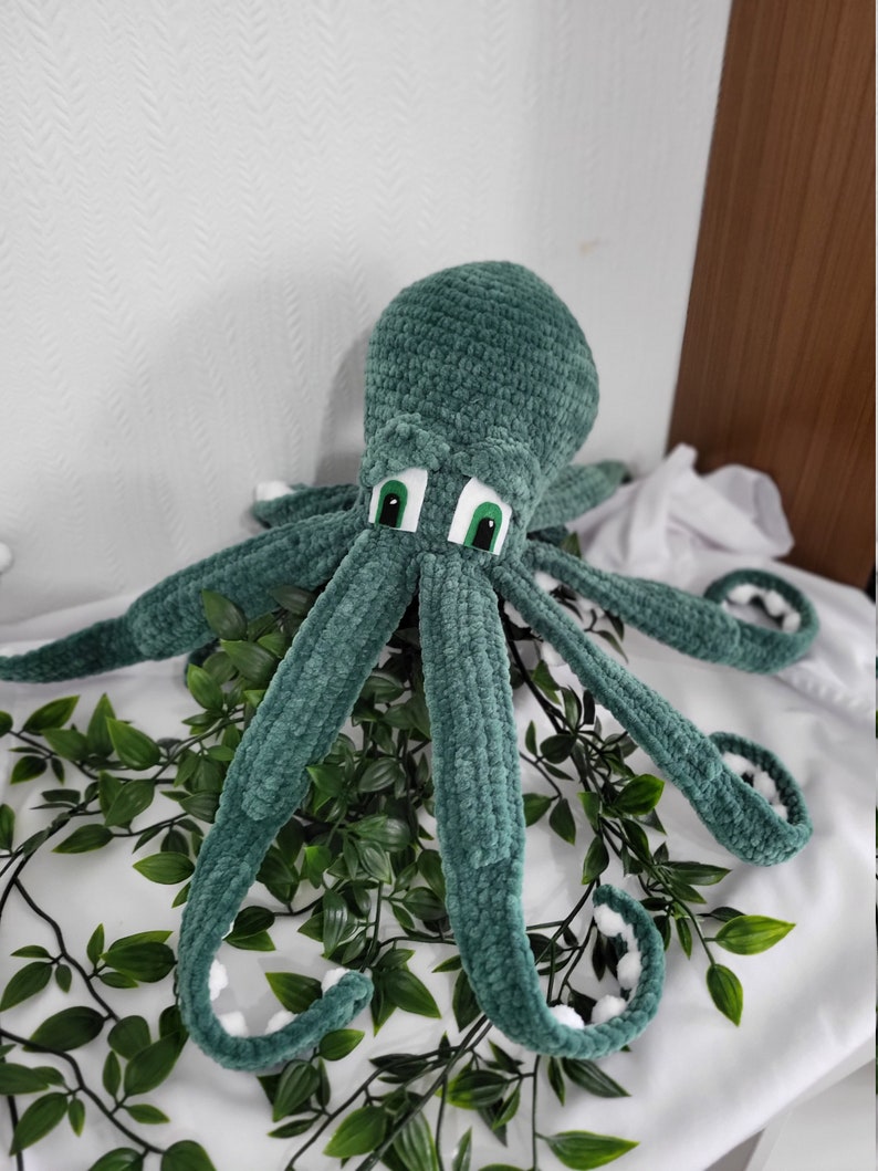 May include: A green crocheted octopus plush toy with white accents on the tentacles and large green eyes. The octopus is sitting on a white surface with green leafy plants.