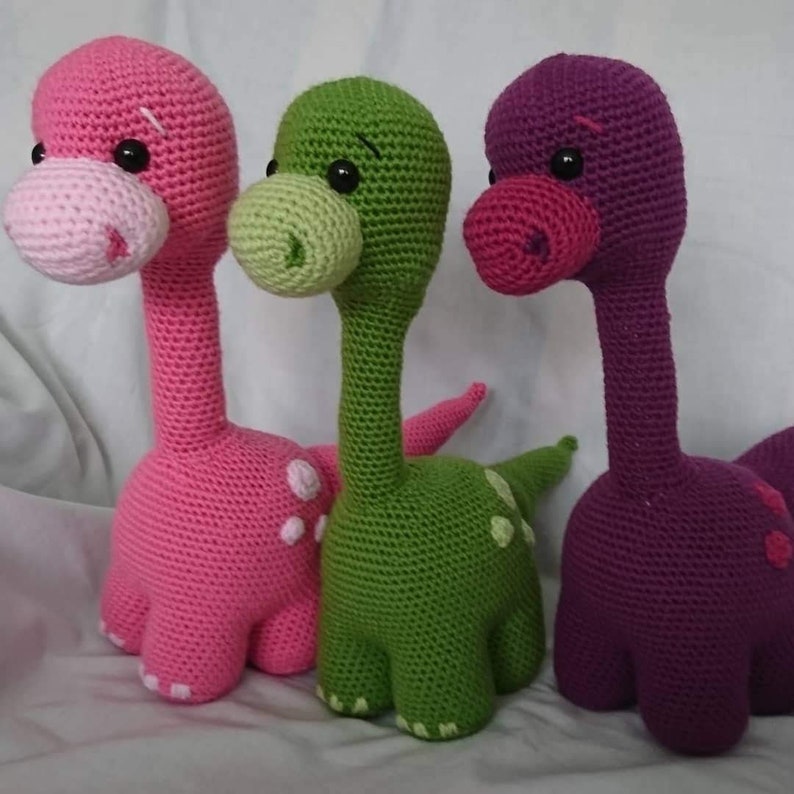 May include: Three crocheted dinosaur toys, one pink, one green, and one purple. Each dinosaur has a long neck, a small head with black eyes, and a tail.