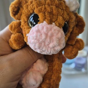 May include: A handmade brown crochet cow plushie with a pink muzzle and white accents. The cow has large black eyes and small, fluffy white horns. The plushie is held in a hand, showcasing its soft, textured appearance.