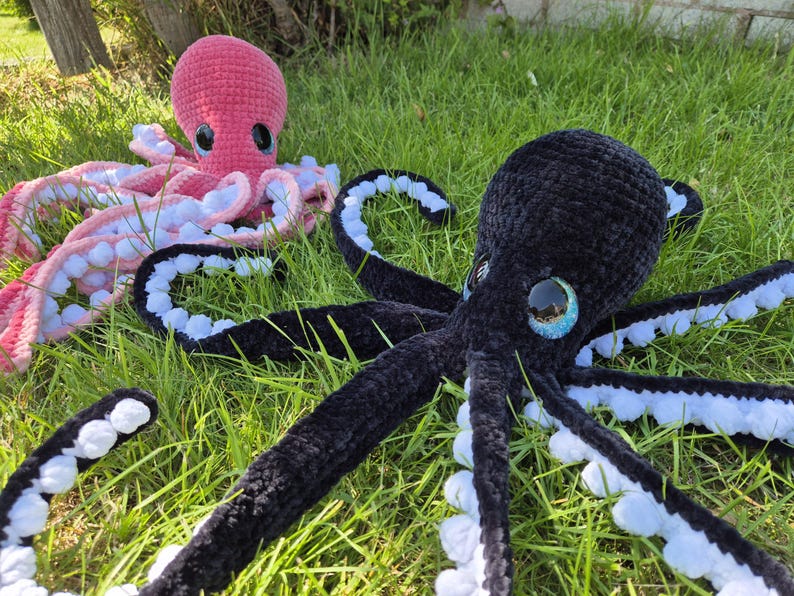 May include: Two crocheted octopus plush toys rest on green grass. One is pink with white and pink tentacles, the other is black with blue and white tentacles. Both have large, round eyes.