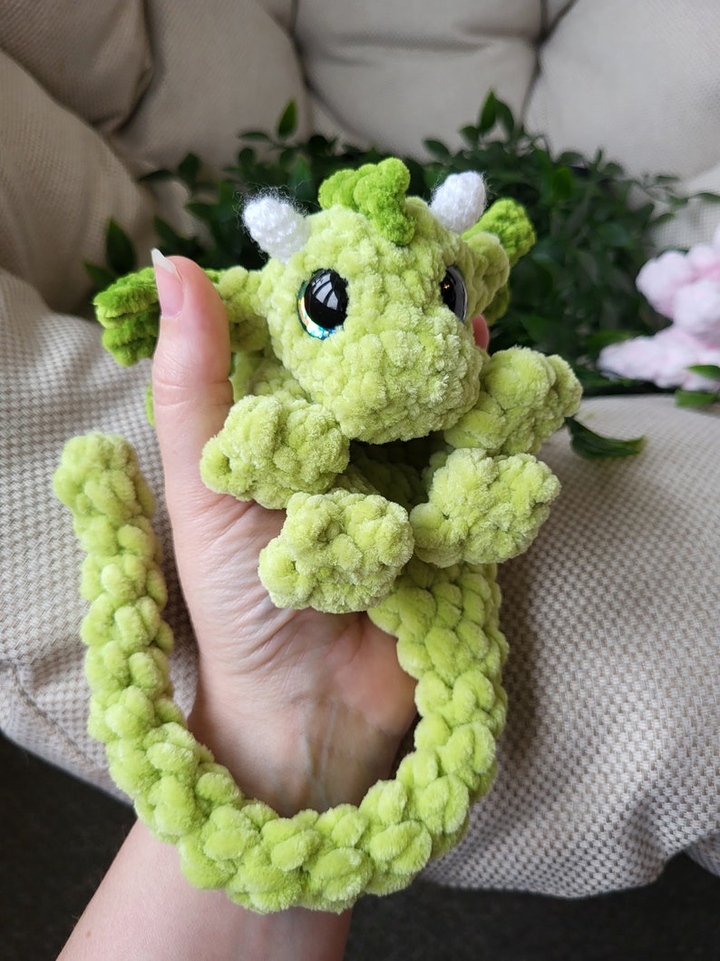 May include: A green crocheted dragon with white horns and sparkly eyes. The dragon has a long, curled tail.