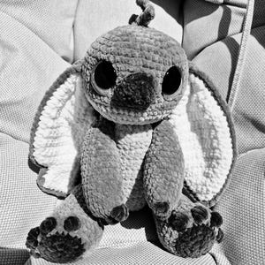 May include: A handmade, grayscale crochet plush toy with large, dark eyes and prominent ears. The plush features a textured, fuzzy appearance and is seated on a textured surface. The toy's feet have detailed paw pads.