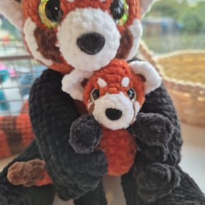 May include: A crocheted red panda plushie, holding a smaller version. The larger toy has a red and white head, large glittery eyes, and a black nose. The body is black with a red and brown tail. The smaller plush has similar features.