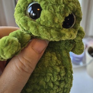 May include: A green crocheted frog plush toy with large, black eyes. The handmade toy has a textured, fuzzy appearance and small, rounded limbs. The frog is a whimsical, soft toy, suitable for children or as a decorative item.