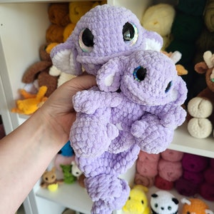 May include: Two crocheted plush toys in the shape of a purple sea creature. The larger creature has a smaller creature nestled in its arms. Both creatures have large, black eyes with iridescent accents.