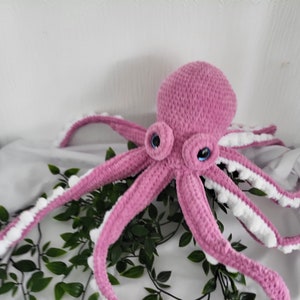 May include: A pink crocheted octopus with white trim on the tentacles and blue eyes. The octopus is sitting on a white surface with green leaves.