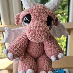 May include: A handmade pink crochet dragon plushie with large black eyes, white horns, and white accents on the wings and feet. The dragon is soft and cuddly, perfect for a child's toy or a decorative item.