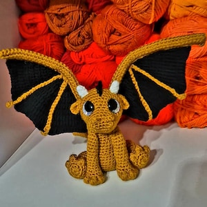 May include: A handmade crochet dragon in shades of gold, black, and yellow. The dragon has large, expressive eyes, small white horns, and black wings with yellow accents. The dragon is sitting with its wings spread, showcasing the intricate details of the crochet work.