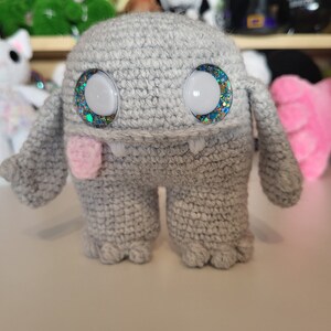 May include: A gray crocheted monster toy with sparkly blue eyes and a pink tongue. The toy has a friendly expression and is standing on a white surface.