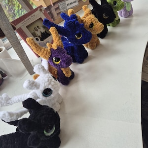 May include: A collection of eight small, plush, crocheted dragon figures in various colors including black, white, purple, blue, tan, and green. Each dragon features large, glossy, dark eyes and textured yarn bodies, arranged in a line on a white surface.