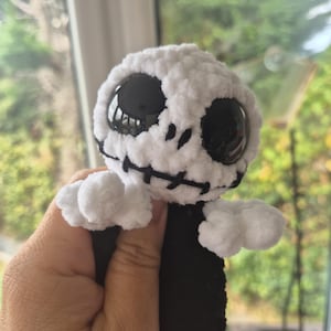 以下が含まれることがあります： 大きな黒いプラスチックの目と黒い刺繍の口を持つ、丸い白い頭のぬいぐるみ。このキャラクターは、柔らかい毛糸で作られた白い質感のある体、小さな白い腕、そして黒い胴体をしており、骸骨のような外観をしています。