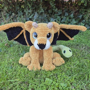 May include: A crocheted plush toy resembling a lion with wings and a snake tail. The lion is brown with black wings and green eyes. The snake tail is green with a green eye.