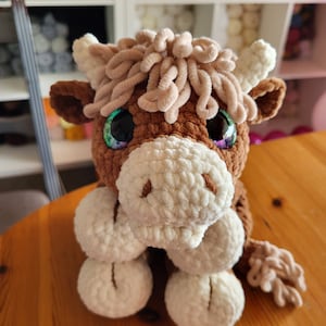 May include: A brown crocheted stuffed animal with white fur on its face and legs. It has large green eyes and a fluffy brown mane. The animal is sitting on a wooden surface.