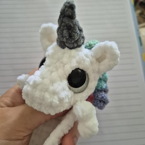 May include: A white crocheted unicorn plush toy featuring a textured yarn body, a dark gray horn, and large black plastic safety eyes. The unicorn has a mane made of soft, curly yarn in shades of light green, light blue, and light purple.