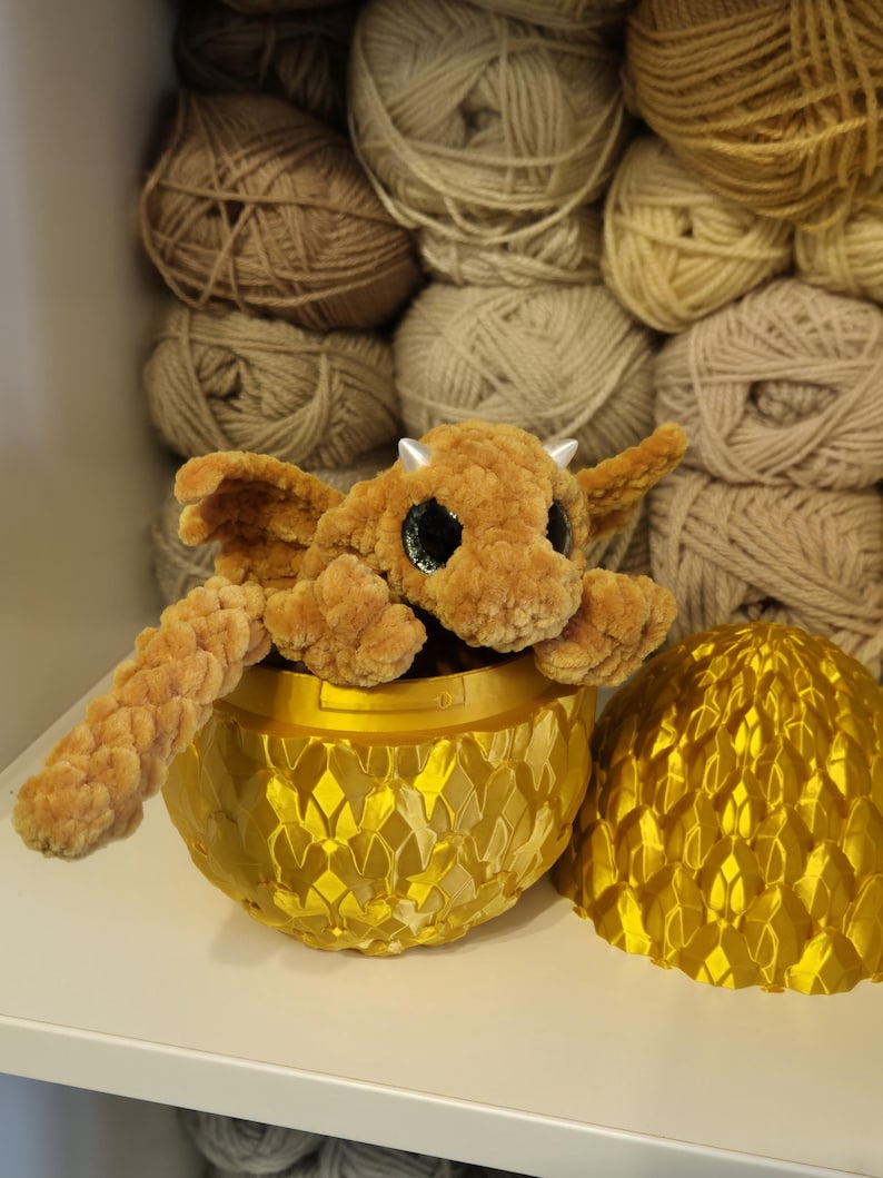 May include: A brown crocheted dragon with large eyes sits inside a gold dragon egg. The egg is open and the top half is beside the dragon.