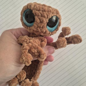 May include: A brown crocheted plush figure with a large, rounded head and a slender body. The figure features two oversized, glossy blue eyes with black pupils, a textured yarn surface, and small, rounded limbs extending from the main body.