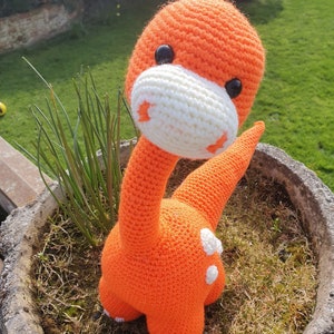 May include: Orange crocheted dinosaur toy with white accents and black eyes. The dinosaur is sitting in a pot with green grass.