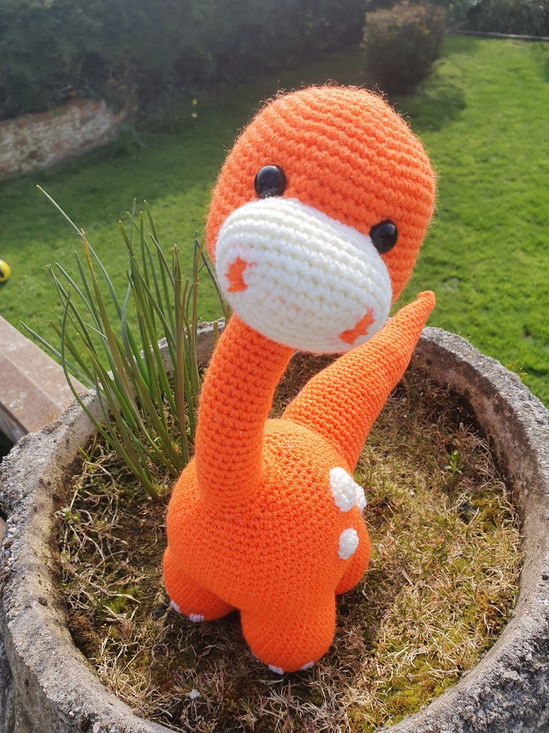 May include: Orange crocheted dinosaur toy with white accents and black eyes. The dinosaur is sitting in a pot with green grass.