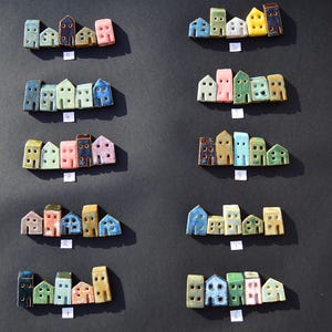 Może przedstawiać: Dziesięć zestawów miniaturowych ceramicznych figurek domków ułożonych w rzędach, każdy zestaw zawiera pięć małych domków w różnych kolorach, w tym niebieskim, zielonym, różowym, żółtym i białym. Każdy zestaw ma małą białą papierową etykietę z numerem od jeden do dziesięć.