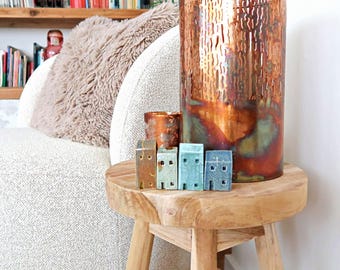 Puede incluir: Una colección de cuatro pequeñas figuras de casas de cerámica en tonos marrón, verde claro y azul, dispuestas sobre un taburete redondo de madera junto a un gran jarrón cilíndrico de metal color cobre texturizado y un portavelas a juego.