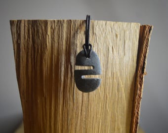 Puede incluir: Un colgante de piedra negra con un diseño tallado cuelga de un cordón negro. El colgante tiene forma ovalada y presenta un diseño simple y abstracto.