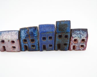 Puede incluir: Una fila de cinco figuras de casas en miniatura de cerámica dispuestas una al lado de la otra. Cada casa presenta un acabado esmaltado único en tonos azules, morados y blancos, con pequeños recortes cuadrados que representan ventanas y puertas.