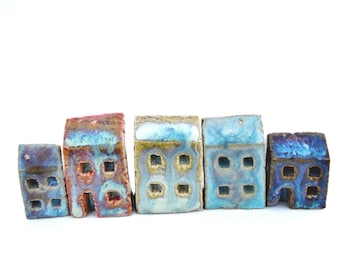 Puede incluir: Un conjunto de cinco pequeñas figuras de casas de cerámica hechas a mano dispuestas en fila. Cada edificio en miniatura presenta un acabado de esmalte reactivo único en tonos azul, marrón y crema, con ventanas cuadradas recortadas y puertas.