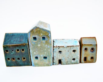 Puede incluir: Un conjunto de cuatro pequeñas esculturas de casas de cerámica dispuestas en fila. Las casas presentan esmaltes variados en tonos azul profundo, verde moteado y blanco pálido, con pequeñas aberturas cuadradas que representan ventanas.