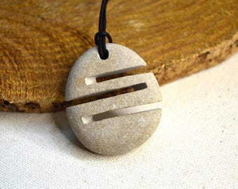 Puede incluir: Un colgante de piedra gris claro con tres secciones horizontales recortadas, suspendido de un cordón negro. El colgante es redondo y tiene un aspecto natural y texturizado. El fondo presenta una superficie de madera.
