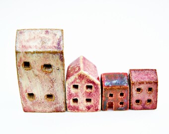 Puede incluir: Un conjunto de cuatro figuras de casas de cerámica de diversas alturas y formas. Cada pieza presenta un esmalte moteado en rosa, morado y crema con pequeños recortes cuadrados que representan ventanas. La estructura más alta es rectangular.