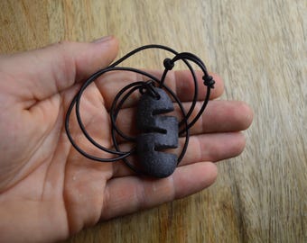 May include: A black stone pendant with a carved design hangs from a black leather cord. The pendant is shaped like a teardrop and has a series of lines carved into it.