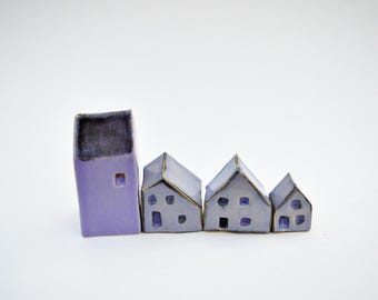 Puede incluir: Un conjunto de cuatro casas de cerámica en miniatura en varios tonos de morado y lavanda. La casa más alta es rectangular con techo plano, mientras que las tres más pequeñas tienen techos inclinados, bordes oscuros y pequeñas ventanas.