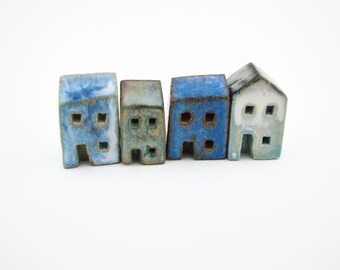 Puede incluir: Cuatro pequeñas figuras de casas de cerámica dispuestas en fila. Las casas presentan esmaltes variados en tonos azul, blanco y gris con recortes cuadrados para ventanas y puertas. Cada pieza tiene una forma y acabado texturizado distinto.