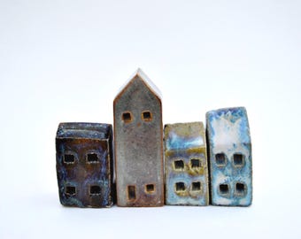 Puede incluir: Un conjunto de cuatro pequeñas figuras de casas de cerámica dispuestas en fila. Las casas presentan esmaltes variados en tonos marrón oscuro, gris, azul y blanco, con ventanas cuadradas recortadas y un techo alto y puntiagudo.