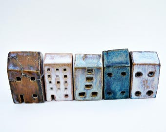 Puede incluir: Cinco pequeñas figuras de casas rectangulares de cerámica dispuestas en fila. Las casas presentan acabados esmaltados variados en tonos marrón, blanco, azul claro y azul oscuro, cada una con pequeñas aberturas cuadradas que simulan ventanas.