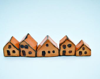 Puede incluir: Una fila de cinco pequeñas figuras de casas de cerámica hechas a mano de diferentes tamaños. Cada casa presenta un esmalte naranja brillante, un techo puntiagudo y ventanas y puertas pintadas a mano en color marrón oscuro.