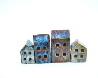 Puede incluir: Un conjunto de cuatro pequeñas figuras de casas de cerámica con acabado esmaltado. Las casas presentan alturas variables y formas rectangulares con ventanas cuadradas recortadas. Las superficies muestran una mezcla de tonos azul profundo, marrón y bronce metálico.
