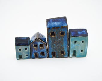 Puede incluir: Un conjunto de cuatro pequeñas figuras de casas de cerámica en varios tonos de azul. Cada pieza presenta un acabado esmaltado con techos azul oscuro y paredes azul claro, incluyendo ventanas y puertas cuadradas dispuestas en una fila.