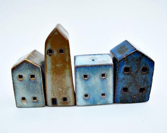 Puede incluir: Cuatro figuras de casas de cerámica dispuestas en fila. La primera es azul claro, la segunda es una estructura marrón alta, la tercera es un bloque rectangular azul pálido y la cuarta es una casa azul oscuro. Cada una tiene pequeñas ventanas cuadradas.