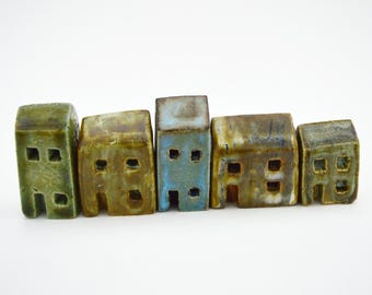 Puede incluir: Un conjunto de cinco pequeñas figuras de casas de cerámica hechas a mano dispuestas en fila. Cada edificio en miniatura presenta un acabado esmaltado en varios tonos de verde, marrón y azul, con ventanas cuadradas y una puerta recortadas.